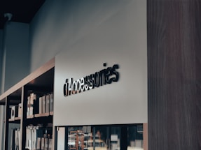 A modern retail store interior with a focus on minimalistic design. Shelves on the left side hold various boxed products, while a sleek, large sign reads 'Accessories' in bold letters. The lighting is soft, creating a calm ambiance.