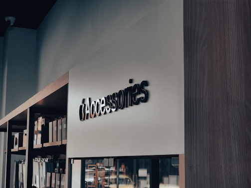 A modern retail store interior with a focus on minimalistic design. Shelves on the left side hold various boxed products, while a sleek, large sign reads 'Accessories' in bold letters. The lighting is soft, creating a calm ambiance.