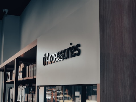 A modern retail store interior with a focus on minimalistic design. Shelves on the left side hold various boxed products, while a sleek, large sign reads 'Accessories' in bold letters. The lighting is soft, creating a calm ambiance.