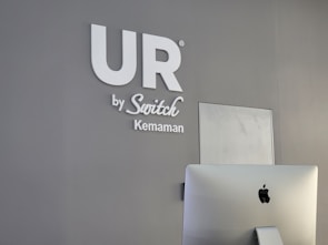 A modern, minimalist office space features a gray wall with the words 'UR by Switch Kemaman' in white letters. An Apple iMac computer is positioned in the foreground, partially visible with the back facing towards the viewer. The overall setting is sleek and professional.
