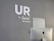 A modern, minimalist office space features a gray wall with the words 'UR by Switch Kemaman' in white letters. An Apple iMac computer is positioned in the foreground, partially visible with the back facing towards the viewer. The overall setting is sleek and professional.