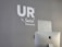 A modern, minimalist office space features a gray wall with the words 'UR by Switch Kemaman' in white letters. An Apple iMac computer is positioned in the foreground, partially visible with the back facing towards the viewer. The overall setting is sleek and professional.