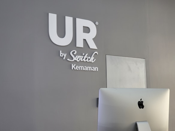 A modern, minimalist office space features a gray wall with the words 'UR by Switch Kemaman' in white letters. An Apple iMac computer is positioned in the foreground, partially visible with the back facing towards the viewer. The overall setting is sleek and professional.