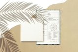 A blank book is placed on top of a detailed form or document with text and diagrams. The background is a beige surface with the shadow of palm leaves cast across the scene, adding a natural and artistic touch.