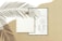 A blank book is placed on top of a detailed form or document with text and diagrams. The background is a beige surface with the shadow of palm leaves cast across the scene, adding a natural and artistic touch.