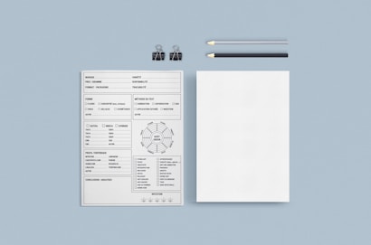 A minimalist layout consisting of a detailed form with multiple sections and options. The form includes checkboxes and text fields on the left. Beside it, there's a blank sheet of paper. Two pencils, one black and one gray, along with two black binder clips, are placed above the papers on a light blue background.