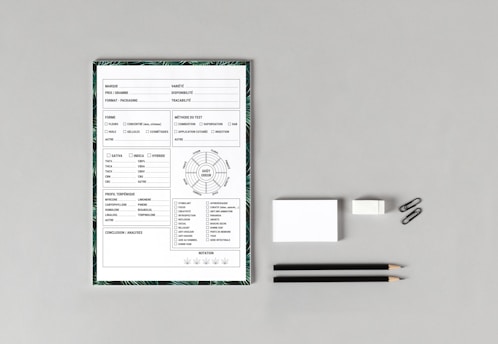 A neatly arranged flat lay featuring a form or checklist with various checkboxes and textual fields, likely related to product specifications or testing. The form is bordered by a leafy pattern. Beside it, there are two black pencils, an eraser, a stack of note cards, and two paper clips, all placed on a gray surface.