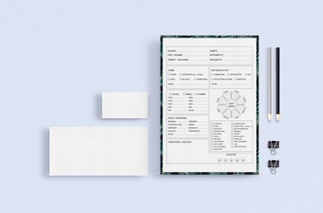 On a light blue background, a stationery set includes a sheet of paper with a form or checklist surrounded by a border with a leaf pattern, two pencils (one black and one light grey) with erasers, two black binder clips, and two blank white cards of different sizes.