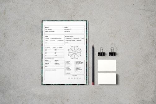 A document with checkboxes and text is placed on a flat, textured gray surface. Next to it, there is a black pencil, two black binder clips, and two small blank cards. The document appears to be some kind of form or template, featuring French text and various sections for input or selection.
