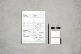 A document with checkboxes and text is placed on a flat, textured gray surface. Next to it, there is a black pencil, two black binder clips, and two small blank cards. The document appears to be some kind of form or template, featuring French text and various sections for input or selection.