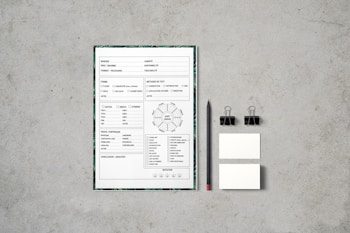 A document with checkboxes and text is placed on a flat, textured gray surface. Next to it, there is a black pencil, two black binder clips, and two small blank cards. The document appears to be some kind of form or template, featuring French text and various sections for input or selection.