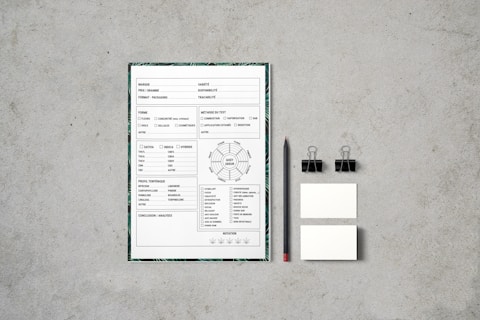 A document with checkboxes and text is placed on a flat, textured gray surface. Next to it, there is a black pencil, two black binder clips, and two small blank cards. The document appears to be some kind of form or template, featuring French text and various sections for input or selection.