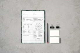 A document with checkboxes and text is placed on a flat, textured gray surface. Next to it, there is a black pencil, two black binder clips, and two small blank cards. The document appears to be some kind of form or template, featuring French text and various sections for input or selection.