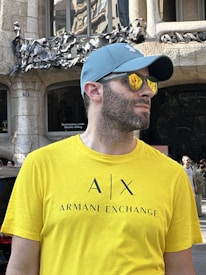 A person wearing a bright yellow Armani Exchange t-shirt and a light blue cap stands in front of a building with intricate stonework and metal decorations. The reflection of the building’s details is visible in the person's yellow-tinted sunglasses.