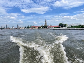 A river scene with a temple on the opposite bank, surrounded by greenery and city buildings. Ripples form in the water, indicating movement from a boat. The sky is partly cloudy with patches of blue.