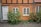 A charming, rustic house with an orange stucco exterior and black timber framing. The building features two small windows with white frames and a white door, along with climbing plants growing up the walls and onto the cobblestone path in front.