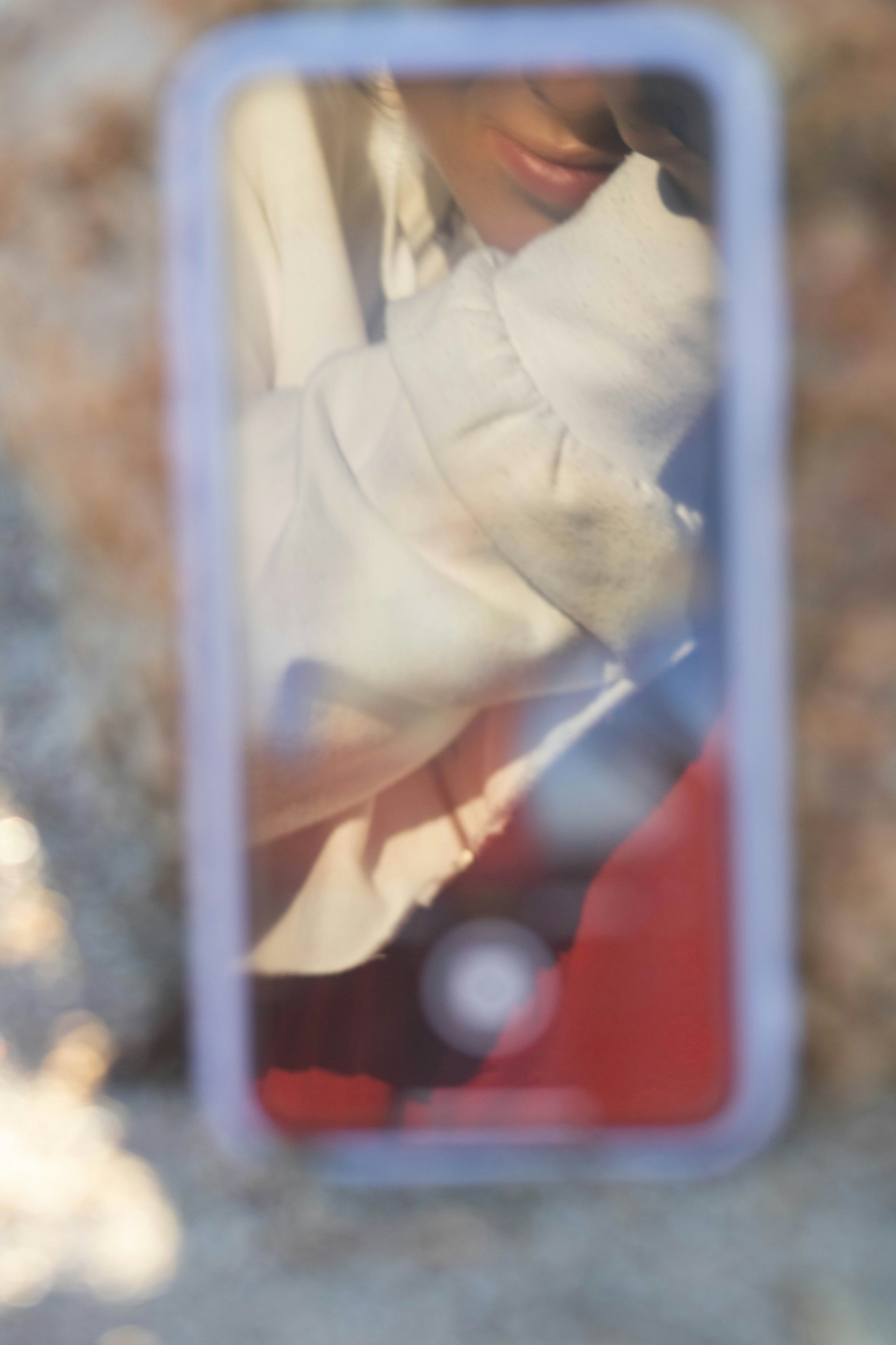A reflective surface, possibly a smartphone, captures the image of a person with their hand near their face, wearing a light-colored sweater. The background is blurred and the image has a soft focus.