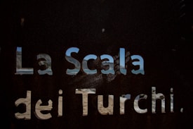 A metal surface with cut-out letters spelling 'La Scala dei Turchi', revealing a scenic view of the sea and sky through the gaps.