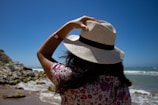 A person stands on the beach facing the ocean, wearing a straw hat and a floral-patterned shirt. Their hand holds onto the hat, shielding from the sun. The sky is a deep blue, and waves gently crash onto the shore. Rocks are visible to the side, and the overall scene conveys a sense of a sunny, breezy day by the sea.