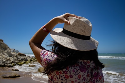 A person stands on the beach facing the ocean, wearing a straw hat and a floral-patterned shirt. Their hand holds onto the hat, shielding from the sun. The sky is a deep blue, and waves gently crash onto the shore. Rocks are visible to the side, and the overall scene conveys a sense of a sunny, breezy day by the sea.