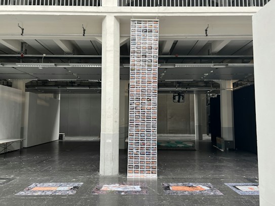 A tall vertical column filled with numerous small images is placed in an industrial-style interior space with concrete walls and exposed ceiling elements. The floor is dark and reflective, with what appear to be art pieces or text on the ground near the column. The setting is open with high ceilings and dim lighting.