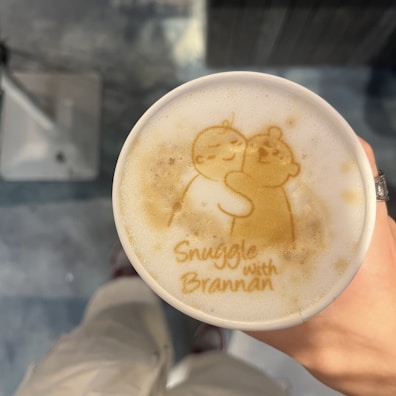A cup of coffee features foam art with a cute illustration of two characters hugging, surrounded by the text 'Snuggle with Brannan.' The cup is being held by a hand wearing a ring, and a blurred background creates a cozy atmosphere.