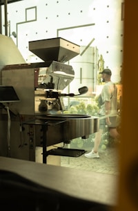 A commercial coffee roasting machine is positioned indoors near a large window, with a glimpse of a person walking outside. The area outside the window features greenery and architectural elements, creating a blend of industrial and natural environments.