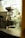 A commercial coffee roasting machine is positioned indoors near a large window, with a glimpse of a person walking outside. The area outside the window features greenery and architectural elements, creating a blend of industrial and natural environments.