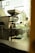A commercial coffee roasting machine is positioned indoors near a large window, with a glimpse of a person walking outside. The area outside the window features greenery and architectural elements, creating a blend of industrial and natural environments.