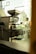 A commercial coffee roasting machine is positioned indoors near a large window, with a glimpse of a person walking outside. The area outside the window features greenery and architectural elements, creating a blend of industrial and natural environments.