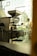 A commercial coffee roasting machine is positioned indoors near a large window, with a glimpse of a person walking outside. The area outside the window features greenery and architectural elements, creating a blend of industrial and natural environments.