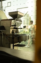 A commercial coffee roasting machine is positioned indoors near a large window, with a glimpse of a person walking outside. The area outside the window features greenery and architectural elements, creating a blend of industrial and natural environments.