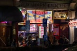 An exterior view of a vibrant restaurant or shop with neon lights featuring the text 'DAVID'S KING OF SPICE'. The area is bustling with people gathered around the entrance. Various colorful signs are displayed in both English and another language. The atmosphere appears lively with a mix of modern and traditional architectural elements.