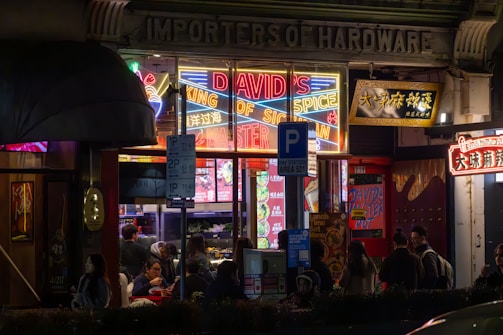 An exterior view of a vibrant restaurant or shop with neon lights featuring the text 'DAVID'S KING OF SPICE'. The area is bustling with people gathered around the entrance. Various colorful signs are displayed in both English and another language. The atmosphere appears lively with a mix of modern and traditional architectural elements.