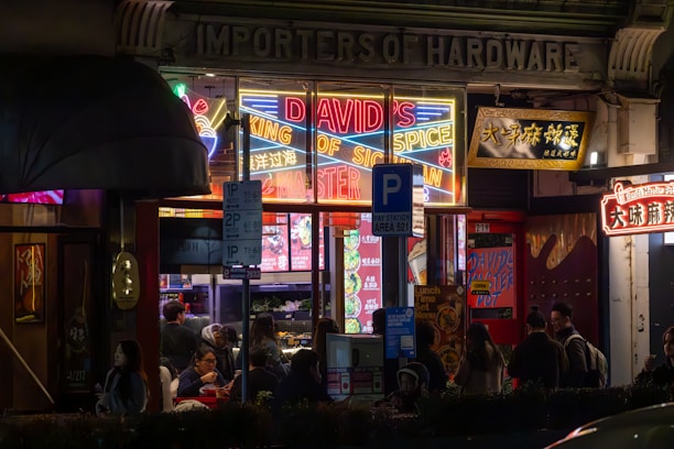 An exterior view of a vibrant restaurant or shop with neon lights featuring the text 'DAVID'S KING OF SPICE'. The area is bustling with people gathered around the entrance. Various colorful signs are displayed in both English and another language. The atmosphere appears lively with a mix of modern and traditional architectural elements.