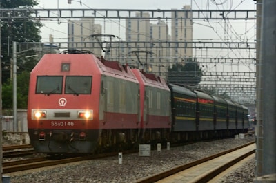 A red and gray electric train moves along the railway tracks with multiple overhead wires and urban buildings in the background. The train is pulling several passenger cars and is surrounded by a complex infrastructure of metal rails and electrical lines.