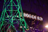 A building with the illuminated sign 'HERALD SUN' in the background, while the forefront features a structure of geometric shapes lit up with bright green lights. The scene is captured at night, adding contrast with the dark sky and purple hues.