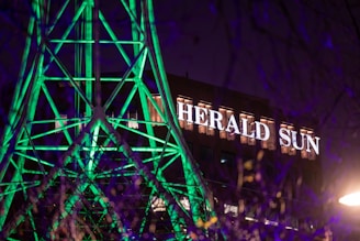 A building with the illuminated sign 'HERALD SUN' in the background, while the forefront features a structure of geometric shapes lit up with bright green lights. The scene is captured at night, adding contrast with the dark sky and purple hues.