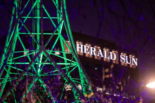 A building with the illuminated sign 'HERALD SUN' in the background, while the forefront features a structure of geometric shapes lit up with bright green lights. The scene is captured at night, adding contrast with the dark sky and purple hues.