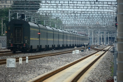 A long passenger train moves along tracks under a complex network of overhead wires, likely for electric trains. The setting appears industrial with visible railway infrastructure such as signals and signs. The background includes trees and urban elements.