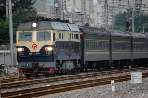 A classic blue and cream-colored locomotive pulling passenger carriages travels along a railway track. Overhead wires suggest it operates on an electrified line. In the background, there are urban buildings and foliage.