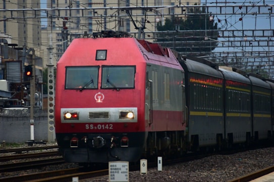 A red locomotive with the number SS9-0142 is pulling a train along a railway track. The background includes urban buildings and overhead electrical wires for the railway. A signal light is visible on the left side of the image, showing a red light.