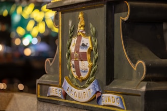 A close-up of a decorative coat of arms on a green and gold structure, featuring detailed engravings with symbols like a ship, a whale, and a lion. The base is inscribed with Latin words and ornamented with leafy designs.