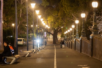 A night-time cityscape with a row of glowing street lamps lining a cobblestone path. On the left, a person sits on a bench, while another couple walks away from the camera on the right side. Lush trees create a canopy overhead, and a vehicle with headlights is parked at the curb.