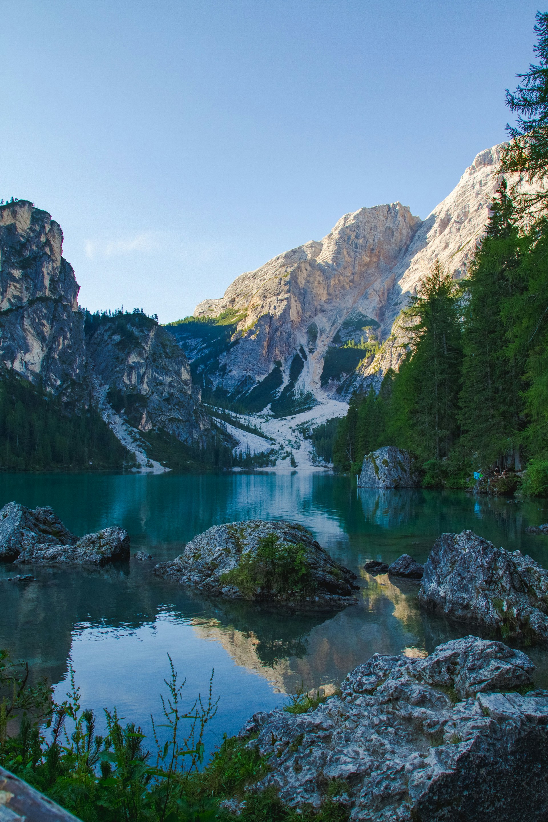 A serene mountain lake reflecting towering pine trees and a clear blue sky at dawn.