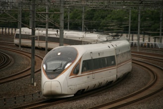 A sleek, modern high-speed train is navigating through curved railway tracks. The train's aerodynamic design features large windows and streamlined bodywork. Overhead power lines are visible, indicating electric propulsion. Lush greenery lines the outside of the track area.