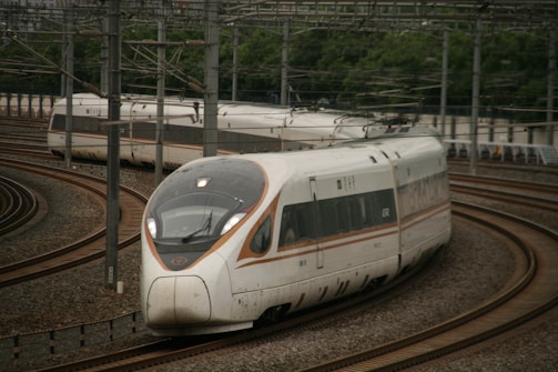 A sleek, modern high-speed train is navigating through curved railway tracks. The train's aerodynamic design features large windows and streamlined bodywork. Overhead power lines are visible, indicating electric propulsion. Lush greenery lines the outside of the track area.