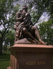 A bronze statue of a seated figure holding a quill pen, with one knee bent. The figure is dressed in historical attire with a flowing coat and rests one hand on a carved stone with inscriptions. The statue sits on a pedestal with the name 'Robert Burns' engraved on it, surrounded by lush green trees and a sunlit park.