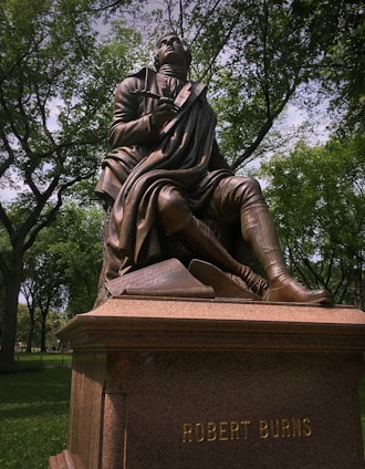A bronze statue of a seated figure holding a quill pen, with one knee bent. The figure is dressed in historical attire with a flowing coat and rests one hand on a carved stone with inscriptions. The statue sits on a pedestal with the name 'Robert Burns' engraved on it, surrounded by lush green trees and a sunlit park.
