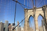 An intricate web of cables and suspension wires frames the stone arches of the Brooklyn Bridge, with the skyline of Manhattan featuring various skyscrapers in the background under a clear blue sky.